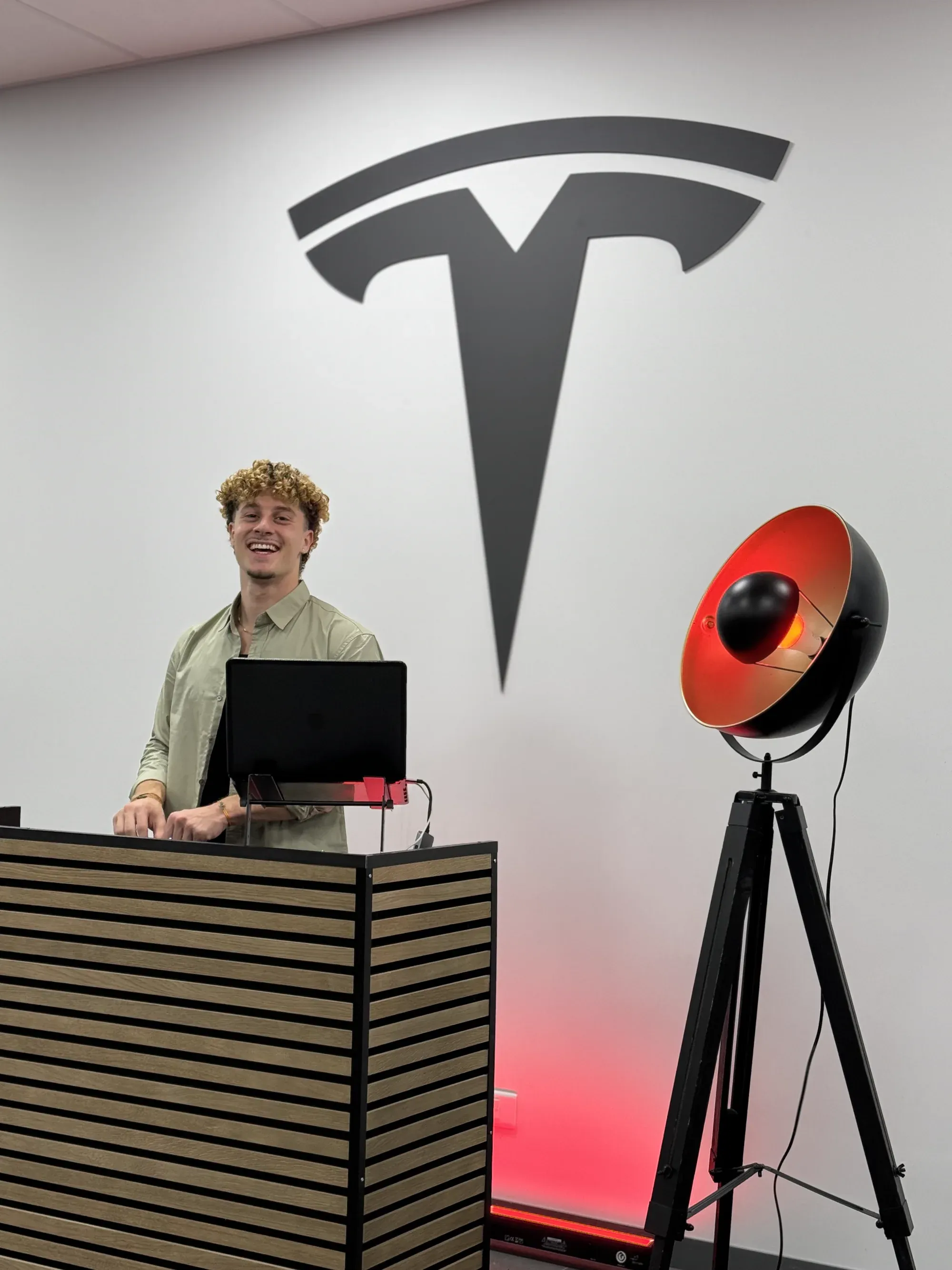 Léo Caussanel, DJ, souriant derrière sa cabine en lattes de bois, sous un grand logo Tesla, à côté d’un projecteur sur trépied