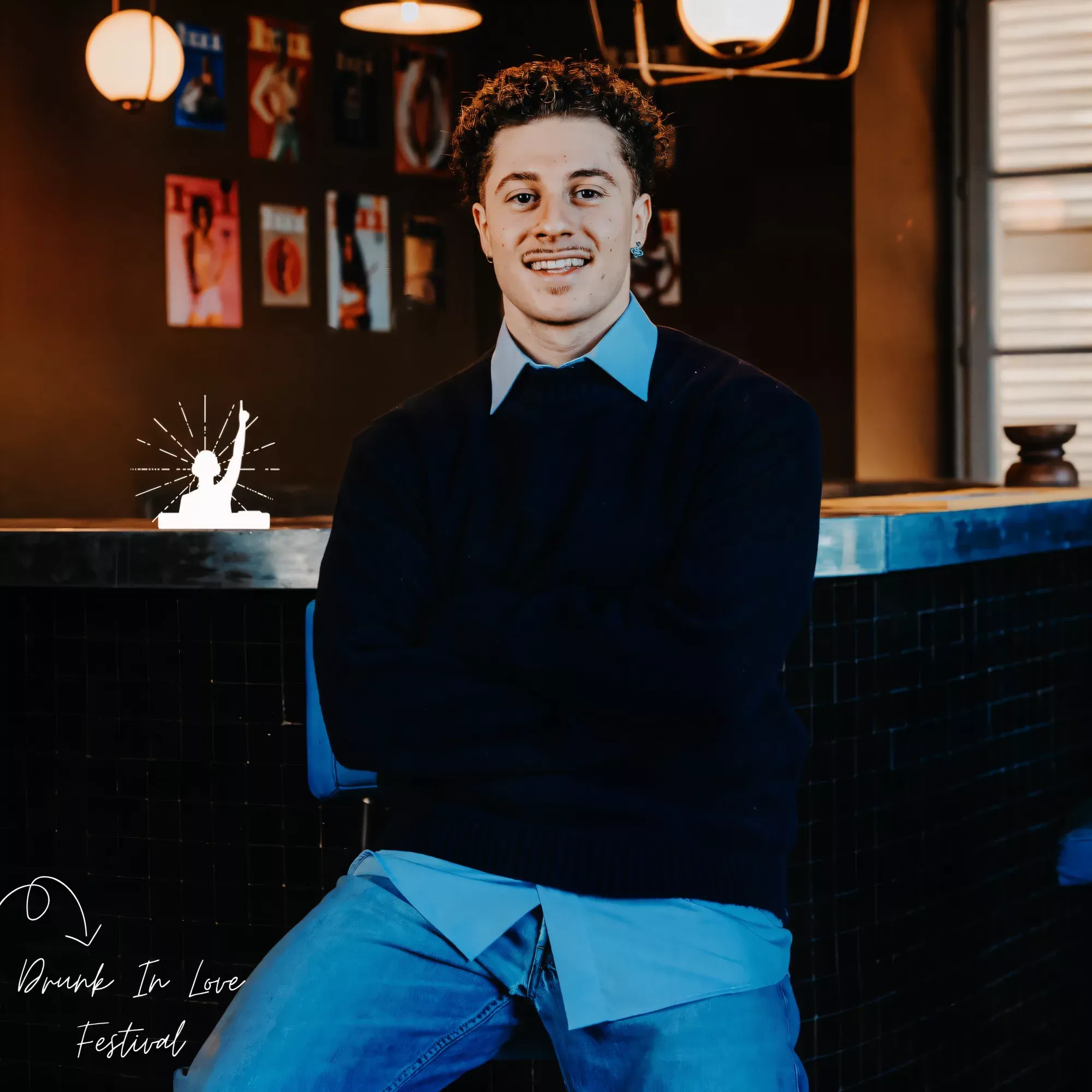 Portrait de Léo Caussanel, DJ, souriant, bras croisés, assis devant le comptoir carrelé d’un bar aux murs couverts d’affiches